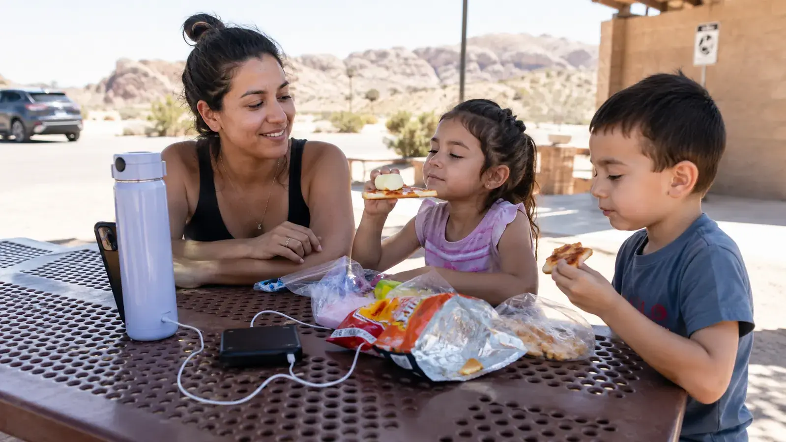 Mom and two kids at a rest stop picnic table, phone plugged into a portable charger while the kids eat snacks